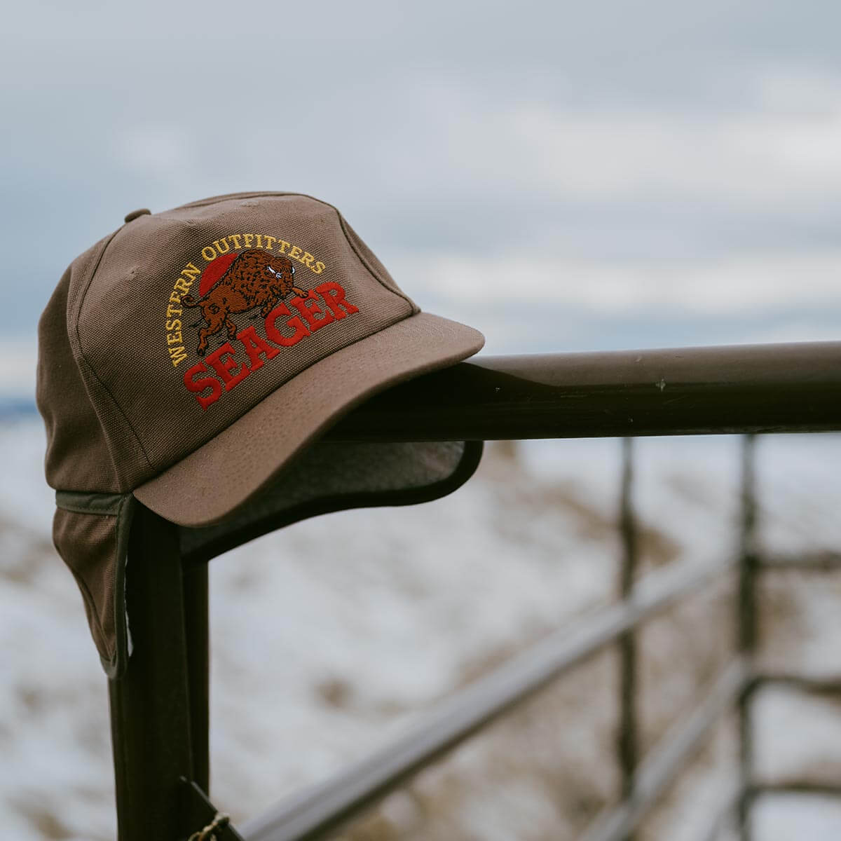 Brown cap with 'Western Outfitters Seager' logo on a metal railing with a blurred natural background