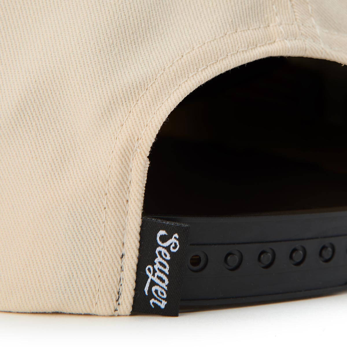 Close-up of a beige cap with a black strap and 'Seater' logo on a white background