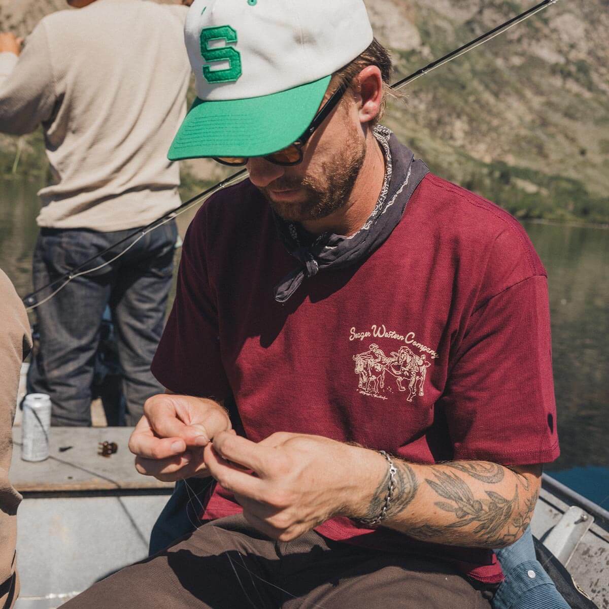 Man in red shirt and green cap sitting on a boat, fishing.