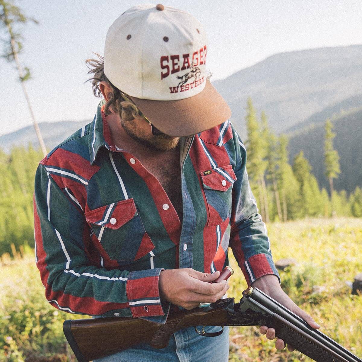 Man holding a shotgun in a field with mountains in the background, wearing a 'Seager' cap.